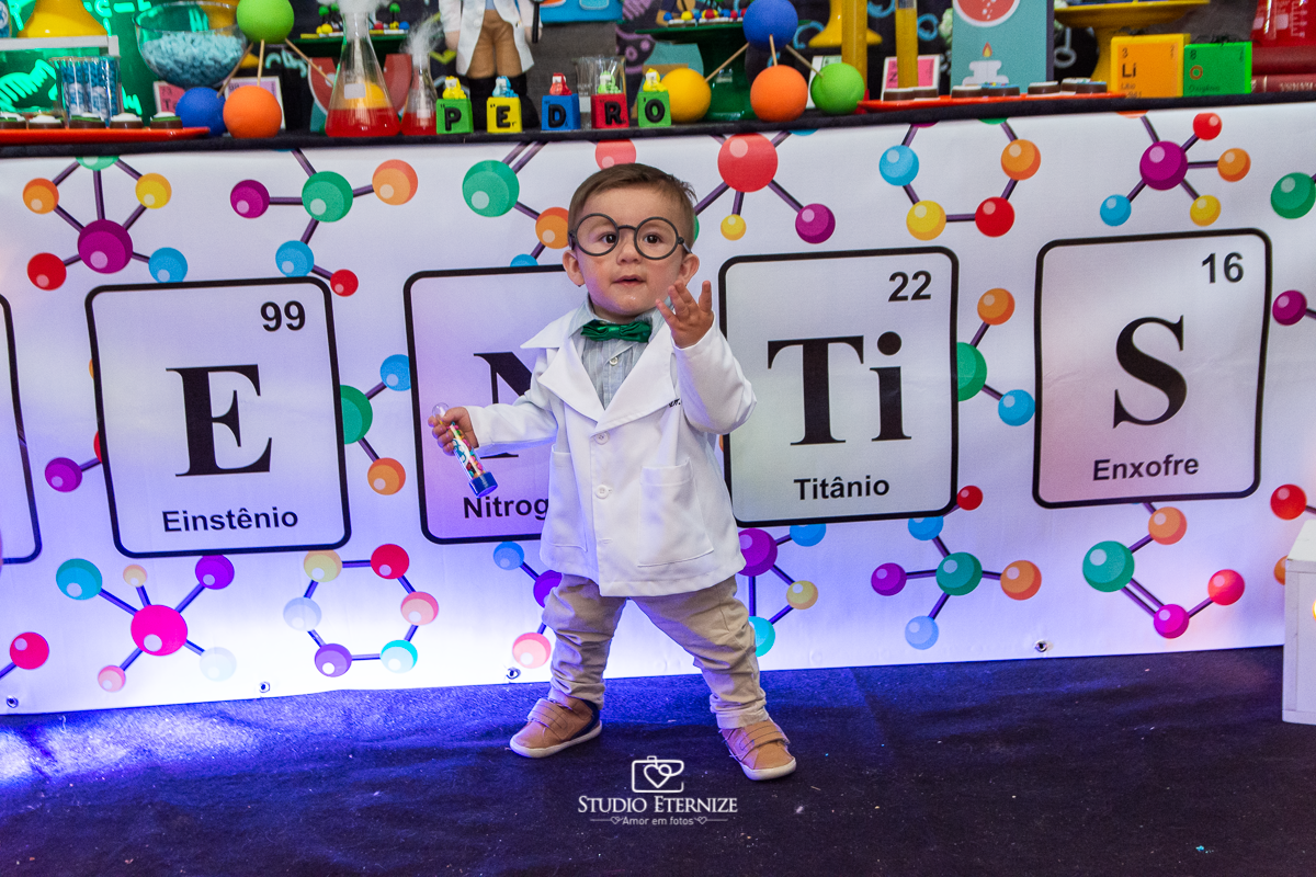 Fotografia de aniversário infantil realizado pelo Studio Eternize em Poços de Caldas. Tema da festa cientista. Aniversariante em pé e de óculos posando para a foto em frente a mesa do bolo.