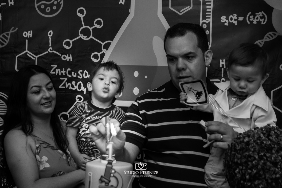 Fotografia de aniversário infantil realizado pelo Studio Eternize em Poços de Caldas. Tema da festa cientista. Irmão do aniversariante no colo de sua mãe soprando a vela enquanto seu pai acende a vela com o aniversariante no colo.