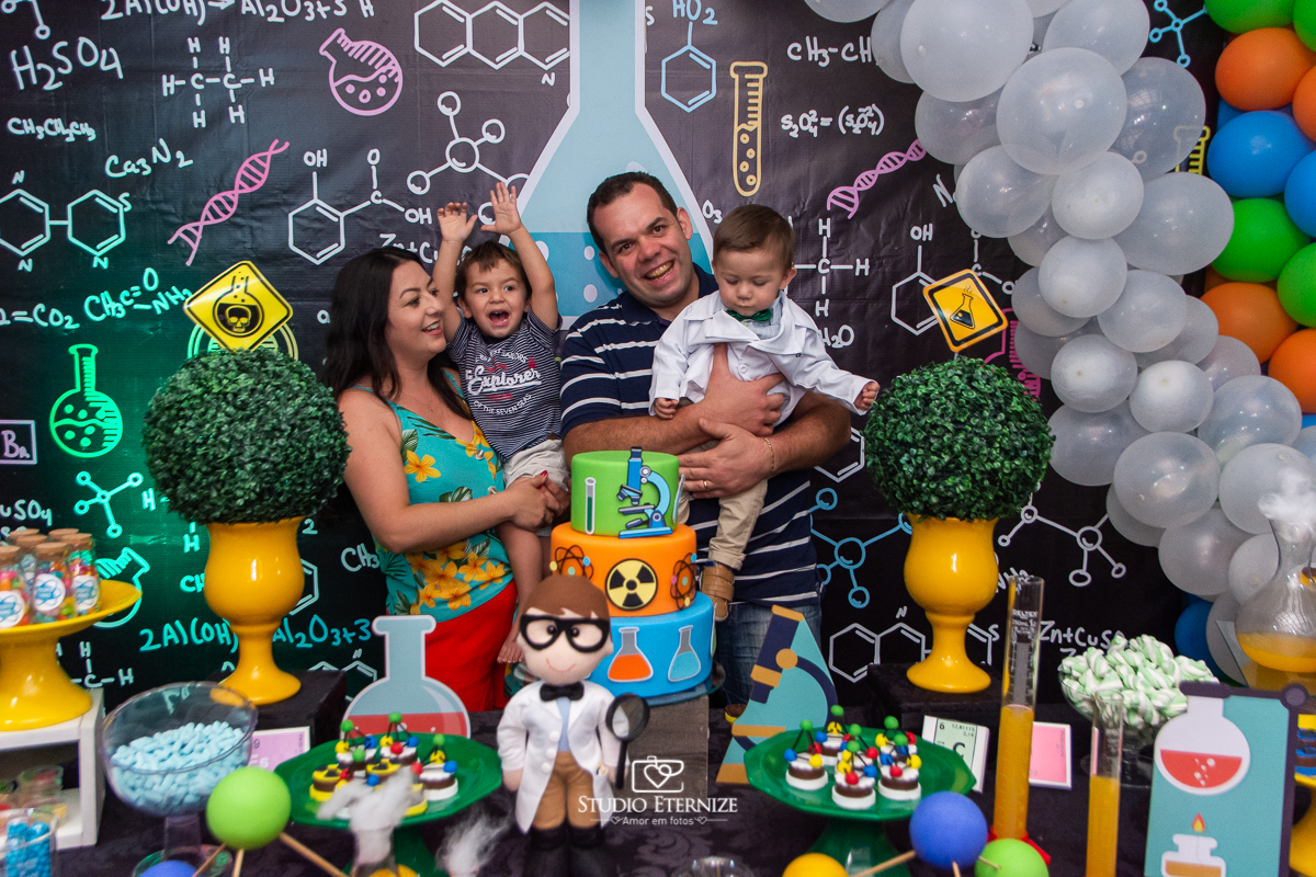 Fotografia de aniversário infantil realizado pelo Studio Eternize em Poços de Caldas. Tema da festa cientista. Irmão do aniversariante vibra no colo da mãe após o parabéns do irmão que está no colo do seu pai.