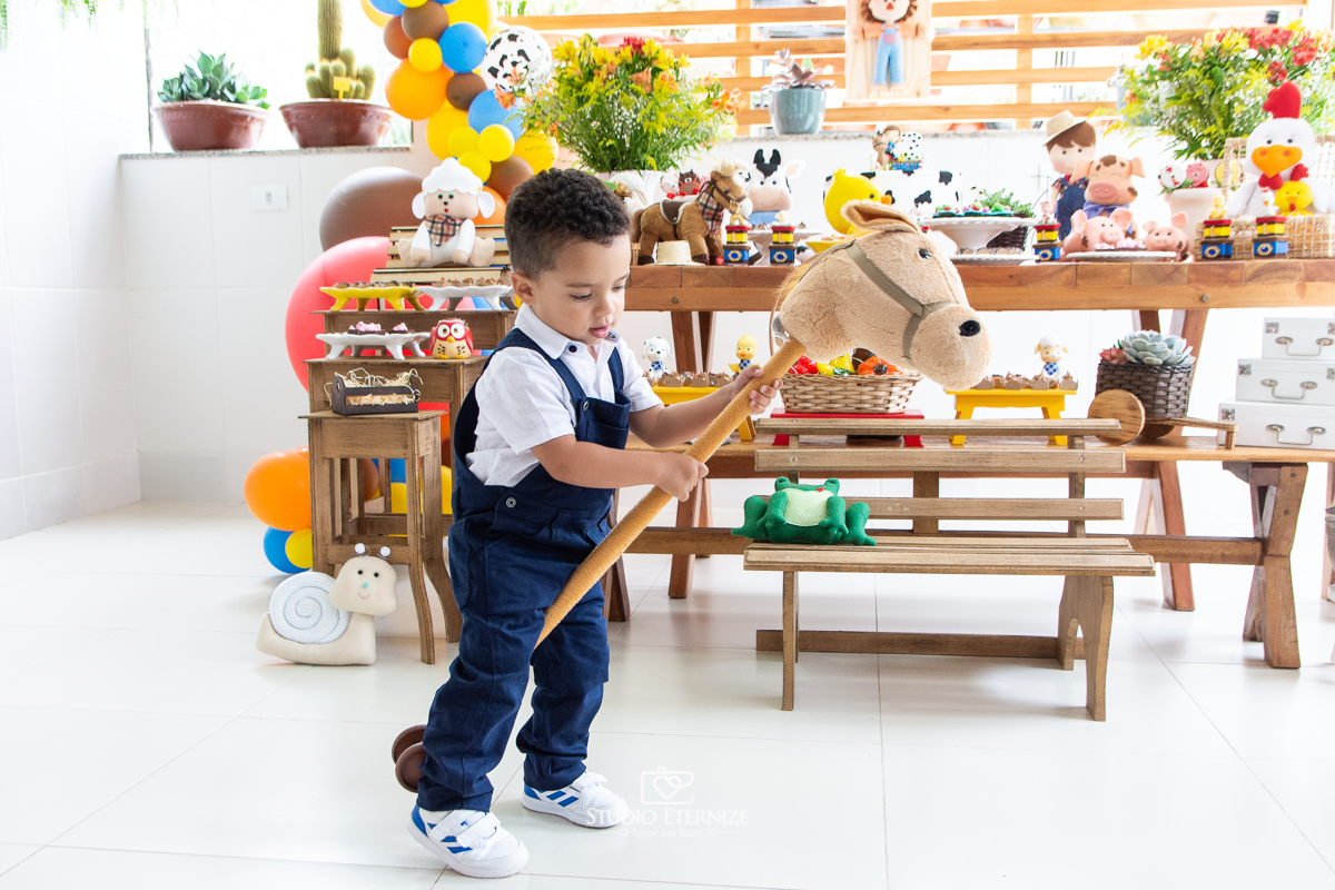 Fotografia de festa infantil de 2 anos registrada pelo Studio Eternize no tema da fazendinha em Poços de Caldas - MG. Aniversariante brinca o cavalinho.