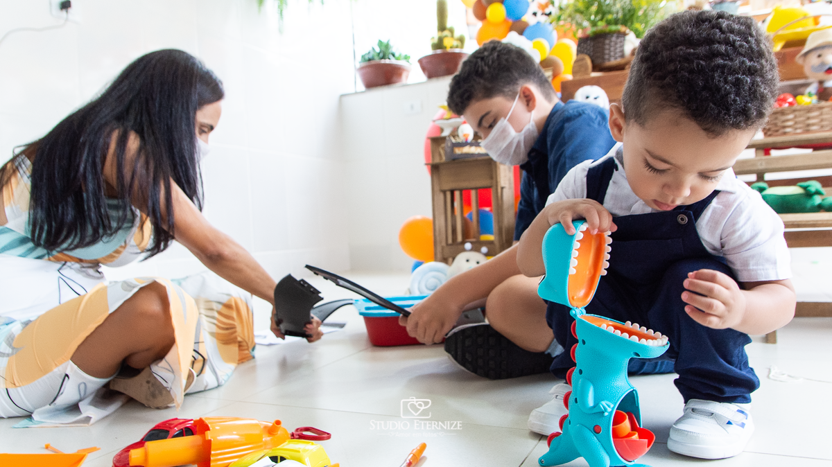 Fotografia de festa infantil de 2 anos registrada pelo Studio Eternize no tema da fazendinha em Poços de Caldas - MG. Aniversariante brinca com o dinossauro que engole as pecinhas enquanto sua mãe e seu irmão montam seu outro presente.