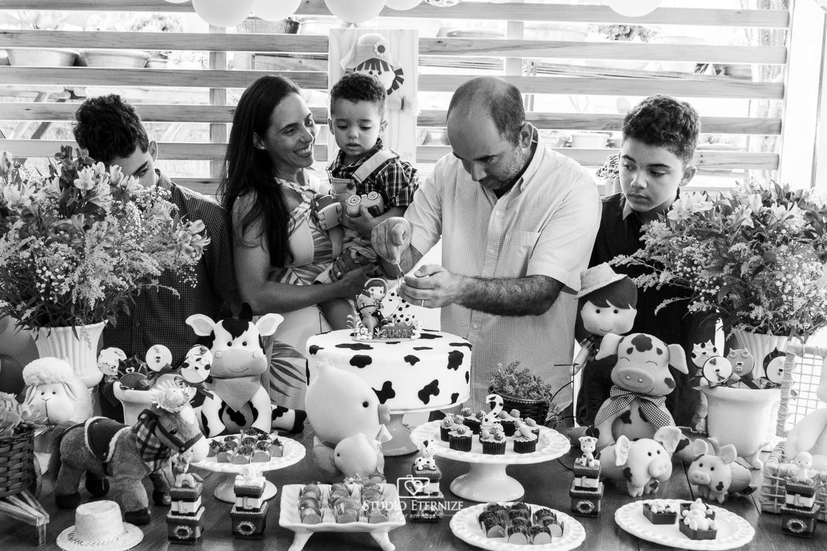 Fotografia de festa infantil de 2 anos registrada pelo Studio Eternize no tema da fazendinha em Poços de Caldas - MG. Aniversariante, seus irmãos e sua mãe observam seu pai acendendo a vela para os parabéns.