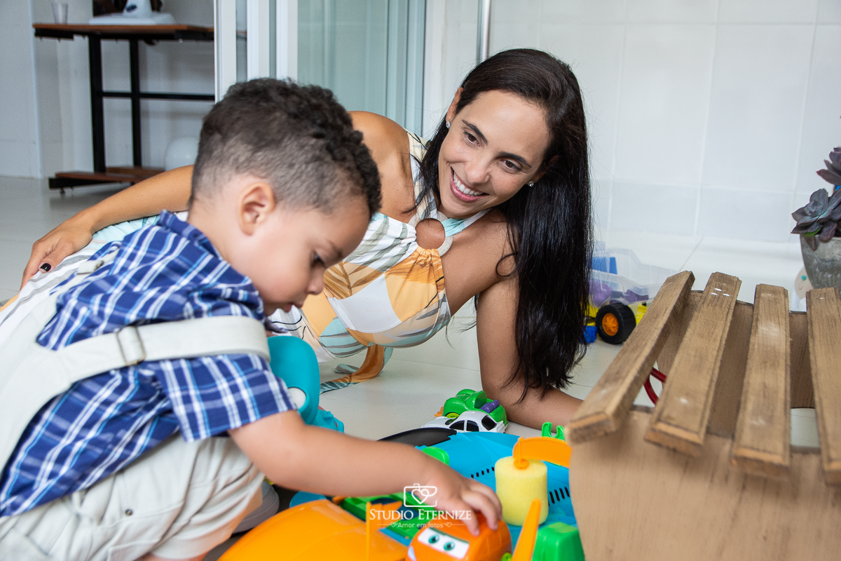 Fotografia de festa infantil de 2 anos registrada pelo Studio Eternize no tema da fazendinha em Poços de Caldas - MG. Aniversariante brinca com presente que ganhou, uma pista de carrinhos enquanto sua mãe o observa encantada.