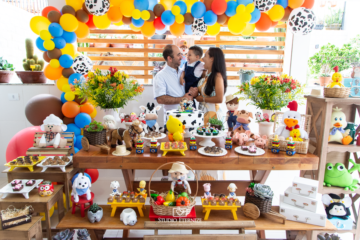 Fotografia de festa infantil de 2 anos registrada pelo Studio Eternize no tema da fazendinha em Poços de Caldas - MG. Aniversariante e seus pais atrás da mesa do bolo, ele olha sorrindo para seu pai.