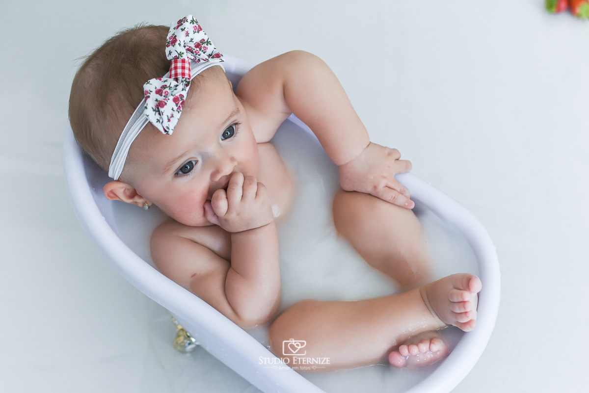 Fotografia de acompanhamento mensal do bebê realizada no Studio Eternize em Poços de Caldas - MG. Bebê menina com 6 meses de idade dentro de uma banheira branca com água branca. Ela morde os dedinhos da mão e está com a perna cruzada.