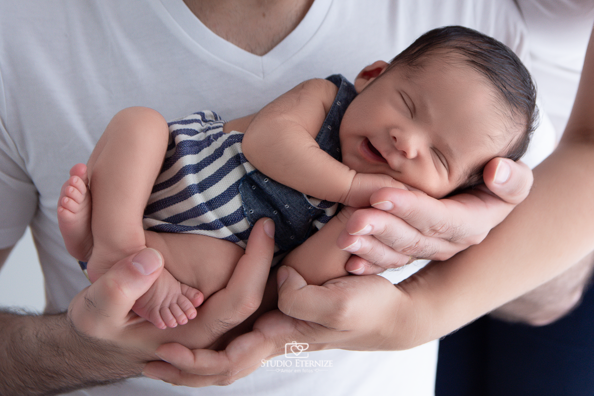 Fotografia newborn menino realizada pelo Studio Eternize em Poços de Caldas - MG. Bebê de roupinha e de ladinho nas mãos do seu pai e da sua mãe.