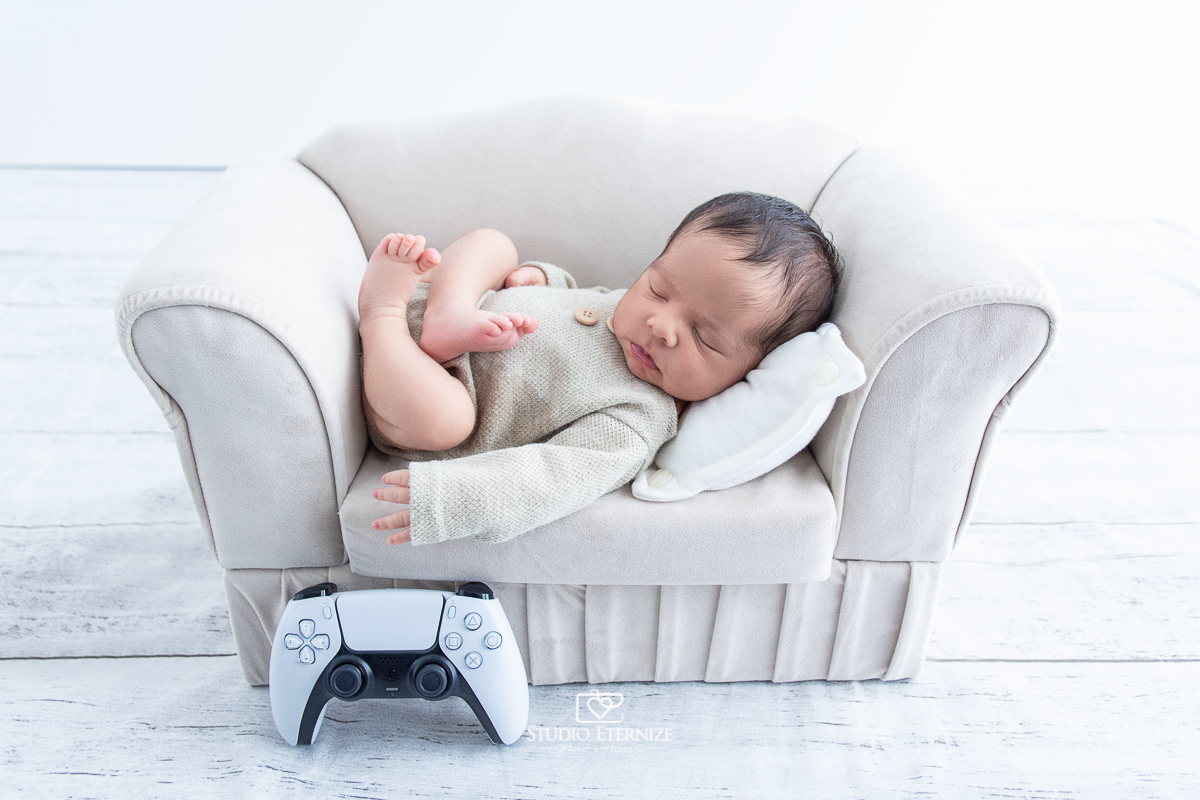 otografia newborn menino realizada pelo Studio Eternize em Poços de Caldas - MG.  Bebê de roupinha com a barriguinha para cima em um sofá pequeno com um controle de videogame ao lado.