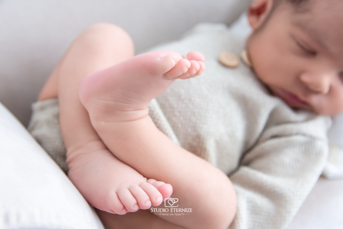 Fotografia newborn menino realizada pelo Studio Eternize em Poços de Caldas - MG.  Bebê de roupinha com a barriguinha para cima em um sofá pequeno, close do seus pezinhos. 