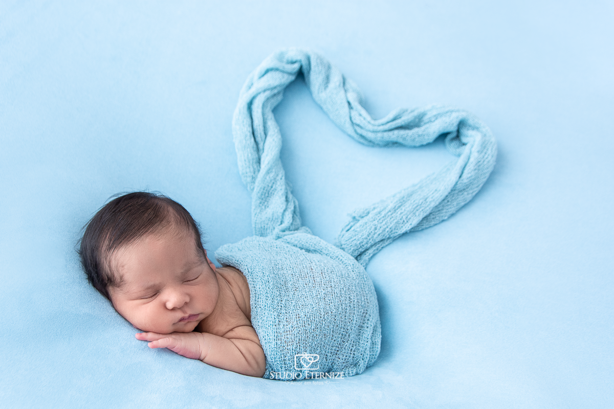 Fotografia newborn menino realizada pelo Studio Eternize em Poços de Caldas - MG. Bebê coberto com wrap que forma um coração na pose de bundinha no puff com uma manta azul.