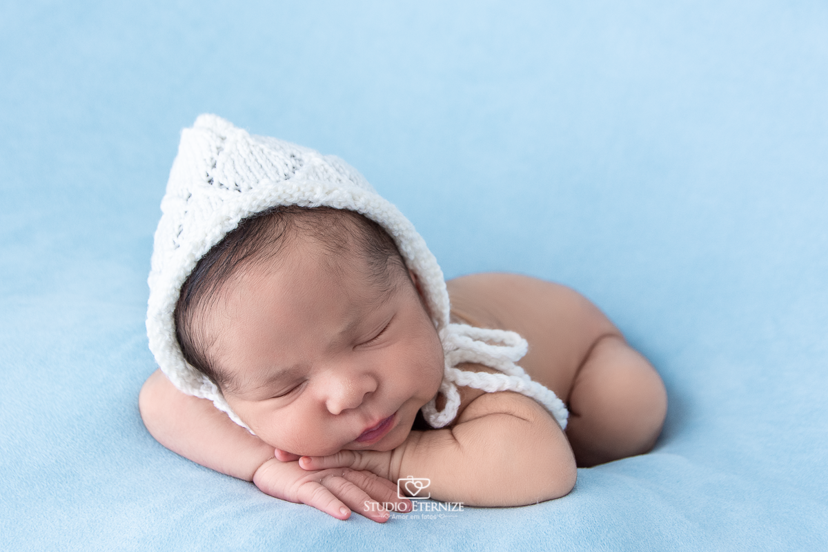 Fotografia newborn menino realizada pelo Studio Eternize em Poços de Caldas - MG. Bebê peladinho com tuquinha branca na pose de bracinho no puff com uma manta azul.