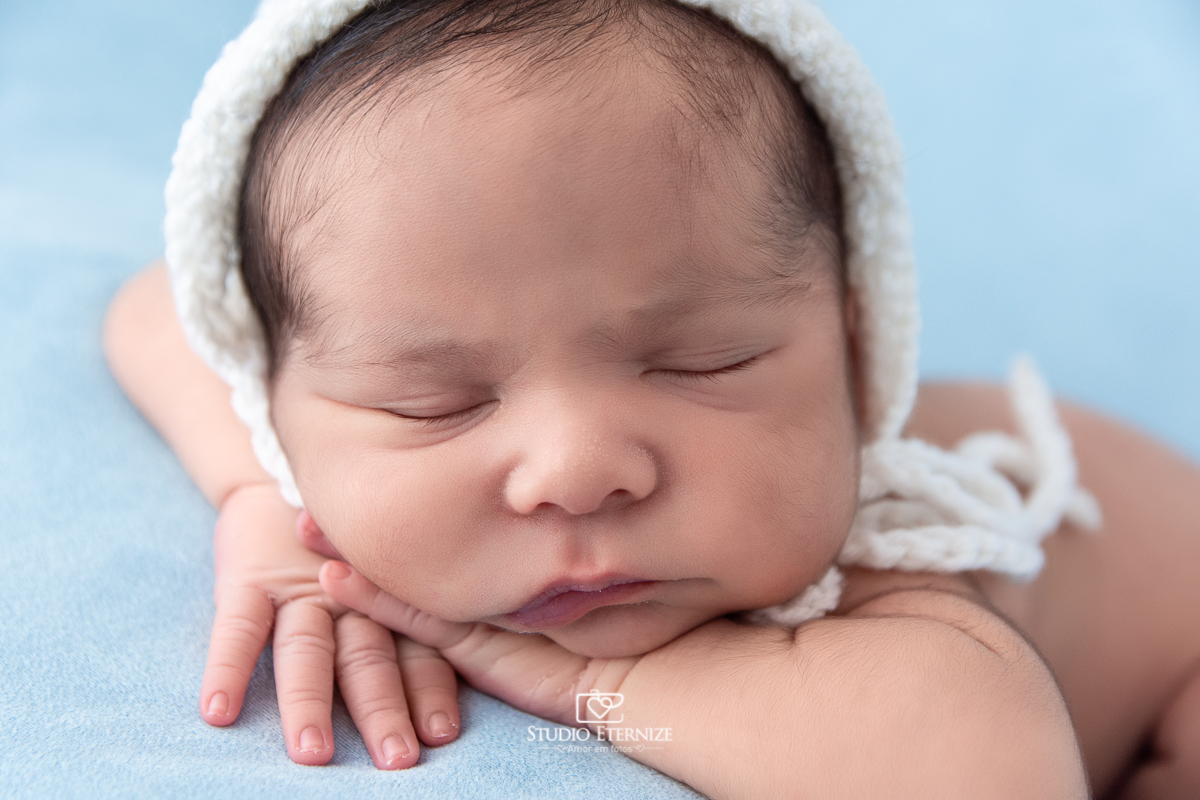 Fotografia newborn menino realizada pelo Studio Eternize em Poços de Caldas - MG. Close do bebê peladinho com toquinha branca na pose de bracinho no puff com uma manta azul.
