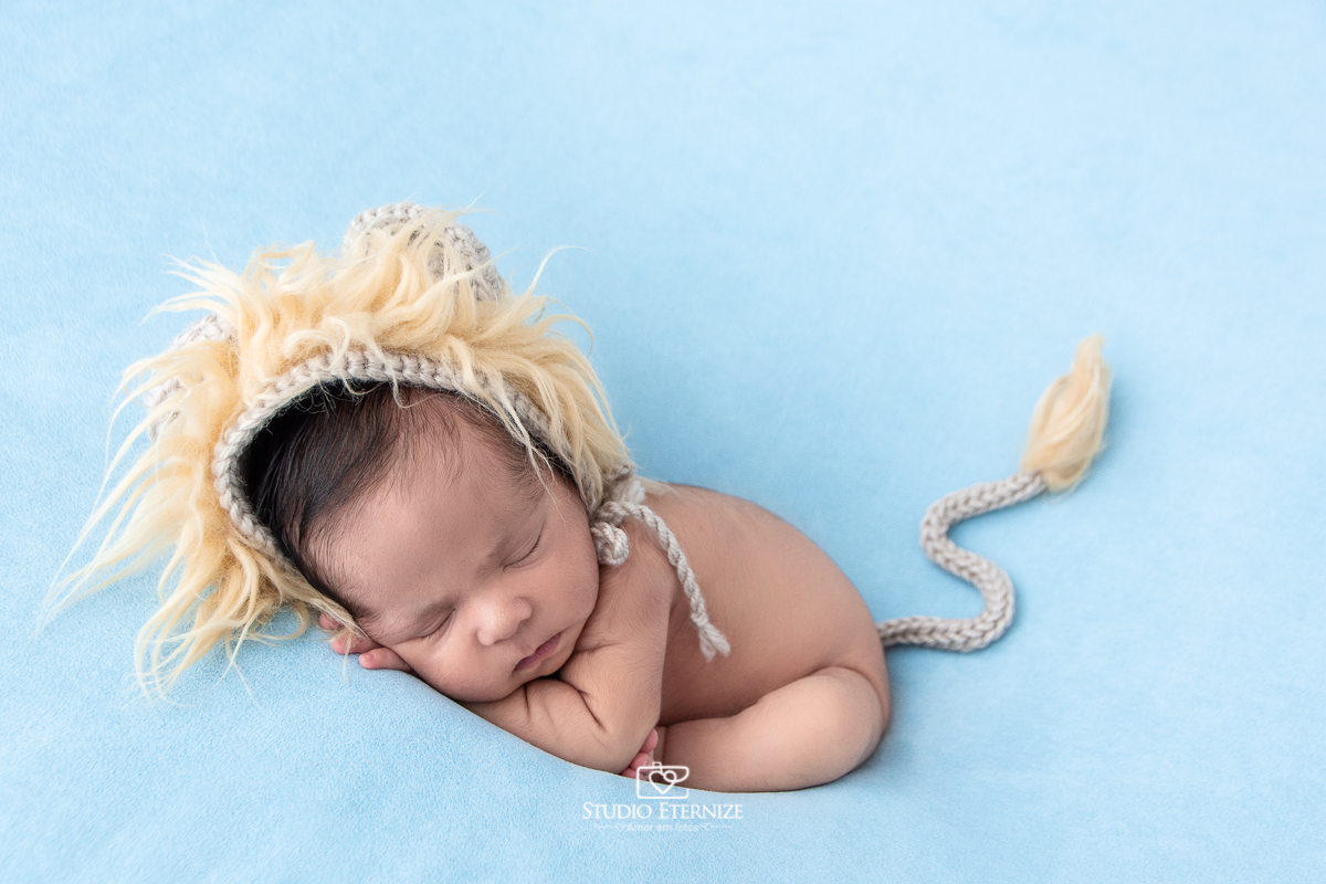 Fotografia newborn menino realizada pelo Studio Eternize em Poços de Caldas - MG. Bebê peladinho com toquinha de leão na pose do colala no puff com uma manta azul.