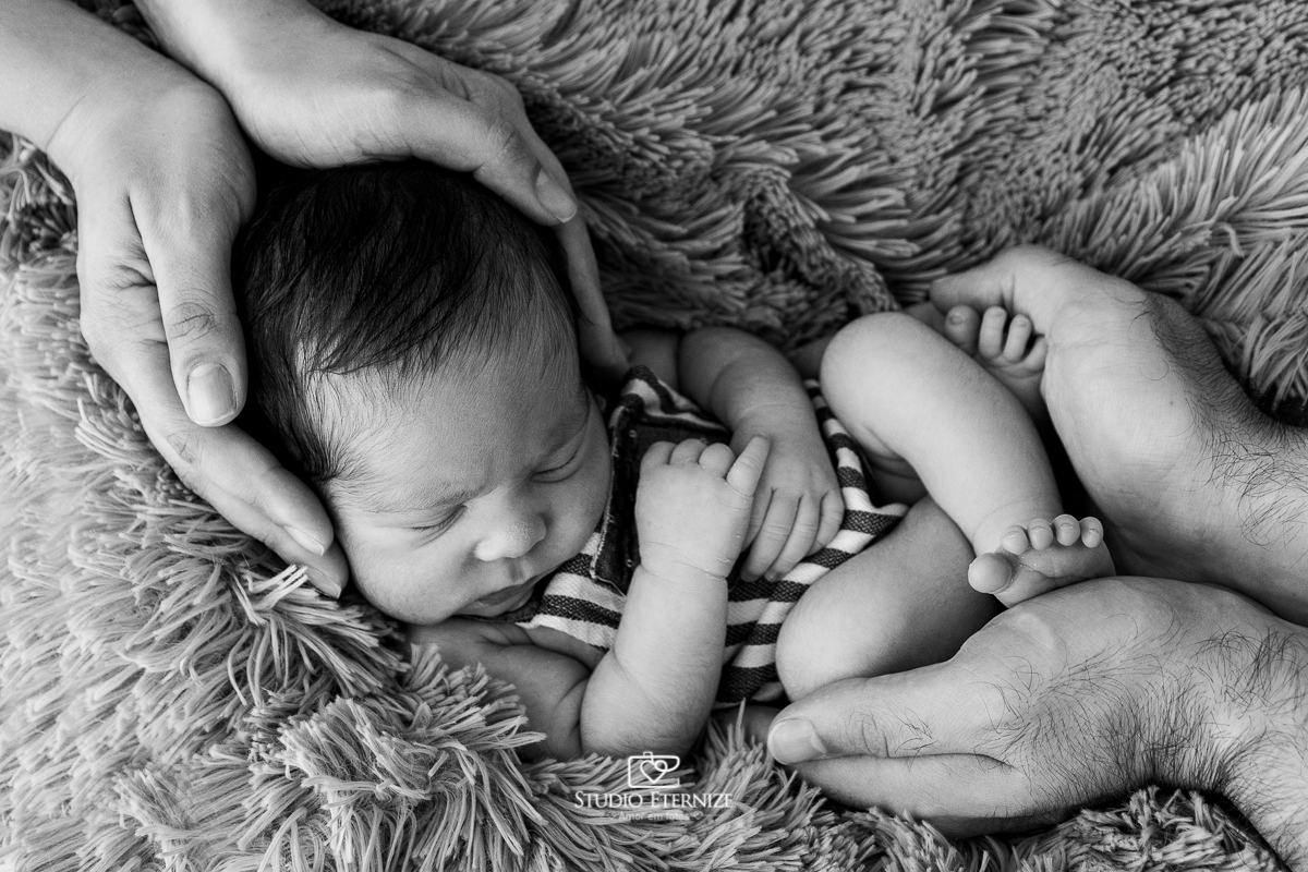 Fotografia newborn menino realizada pelo Studio Eternize em Poços de Caldas - MG. Bebê com roupinha de barriguinha para cima envolto das mãos dos seus pais. Fotografia em preto e branco.