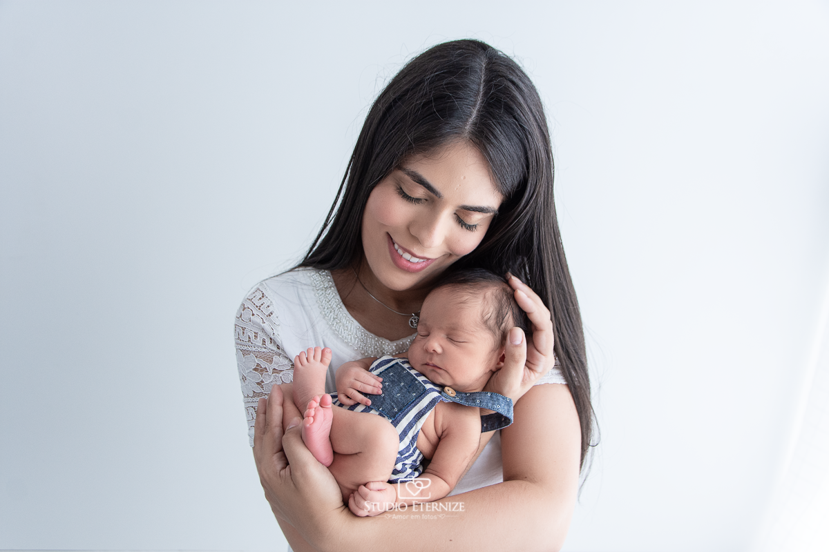 Fotografia newborn menino realizada pelo Studio Eternize em Poços de Caldas - MG. Bebê de roupinha com a barriguinha para cima colo de sua mãe.