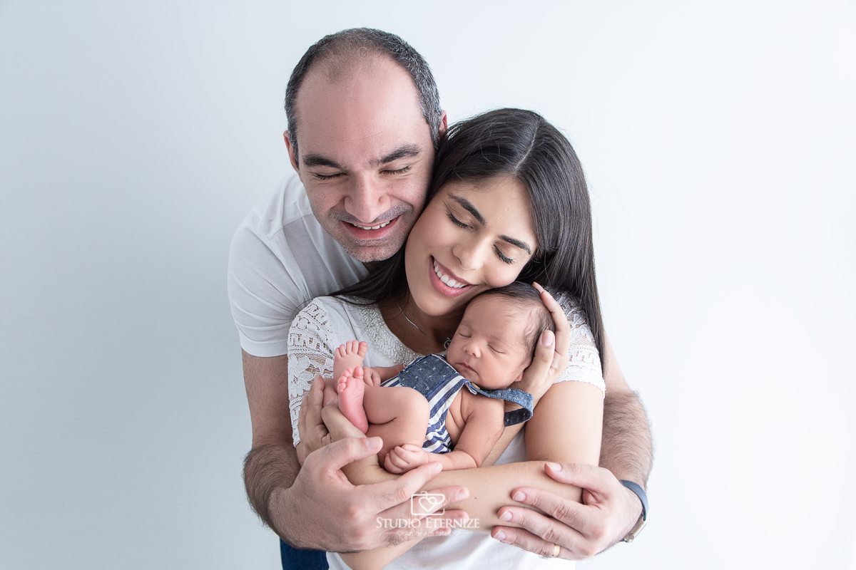 Fotografia newborn menino realizada pelo Studio Eternize em Poços de Caldas - MG. Bebê de roupinha com a barriguinha para cima colo de sua mãe com seu pai abraçando-a.