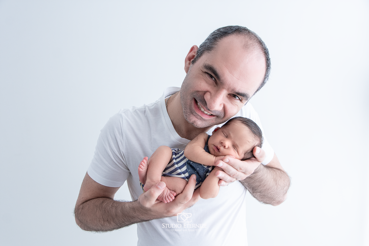 Fotografia newborn menino realizada pelo Studio Eternize em Poços de Caldas - MG. Bebê de roupinha e de ladinho nas mãos do seu pai.