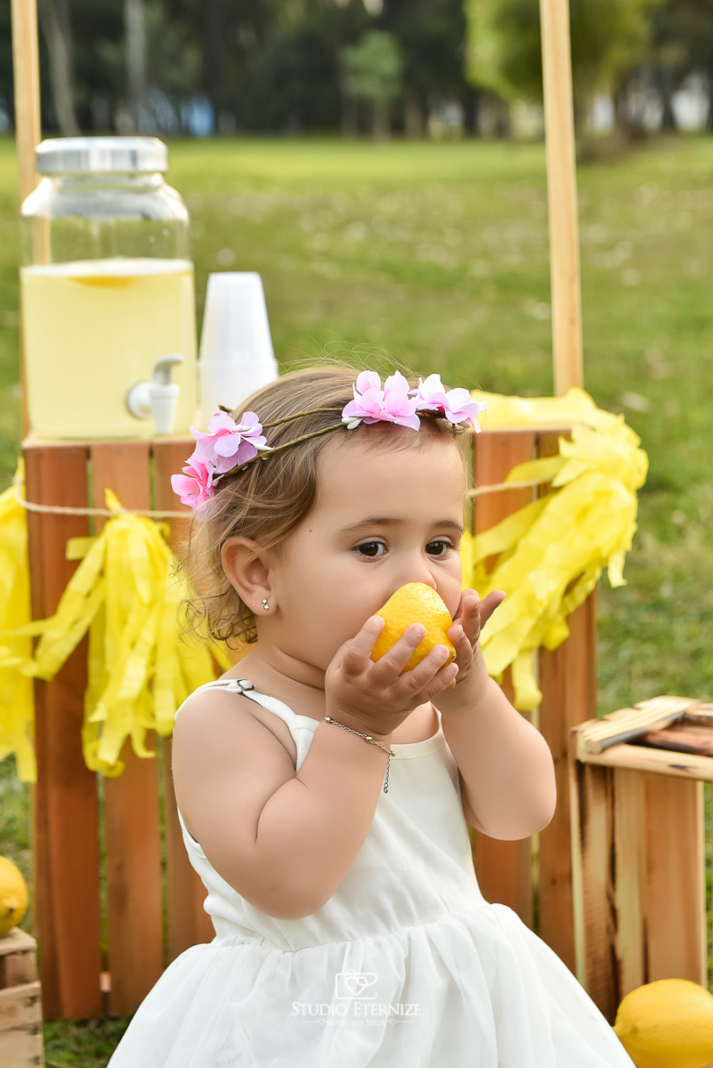 Sessão fotográfica infantil externa com cenário da barraca da limonada. Close da criança sentada na frente da  barraca da limonada experimentando um limão siciliano.