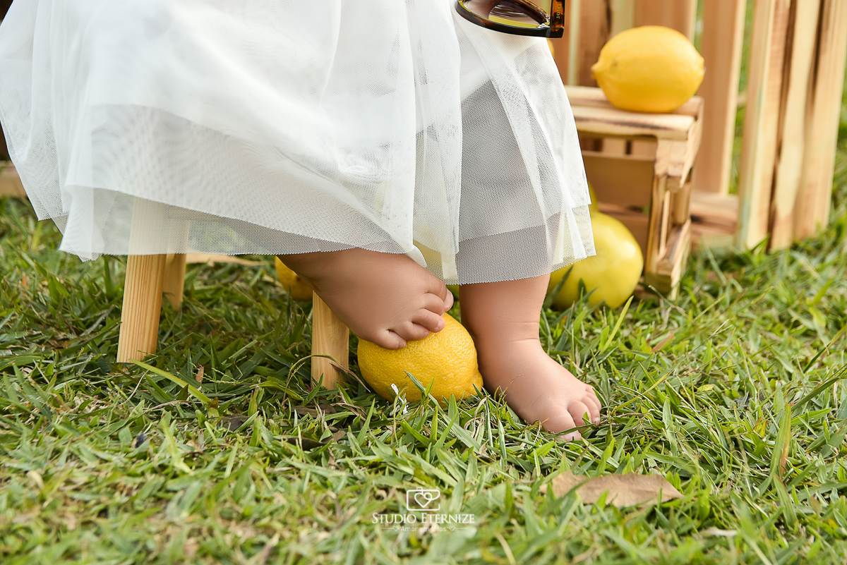 Sessão fotográfica infantil externa com cenário da barraca da limonada. Close dos pés da criança, um pé está apoiado em um limão.