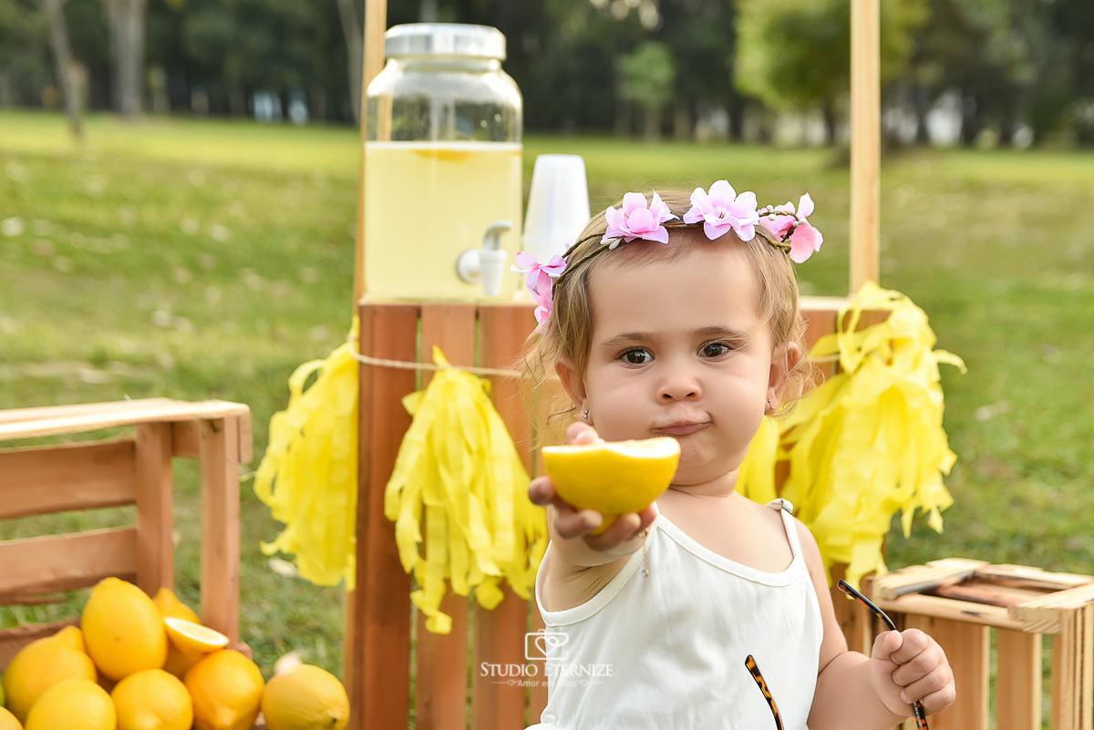 Sessão fotográfica infantil externa com cenário da barraca da limonada. Close da criança sentada na frente da  barraca da limonada oferecendo para a câmera um limão siciliano.