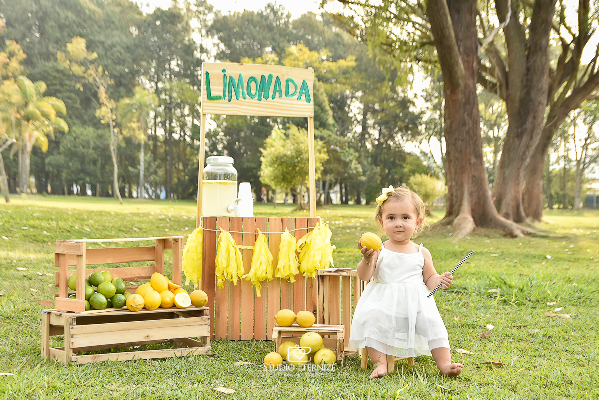 Sessão fotográfica infantil externa com cenário da barraca da limonada. A criança pousa para foto sentada em um banquinho segurando um limão.