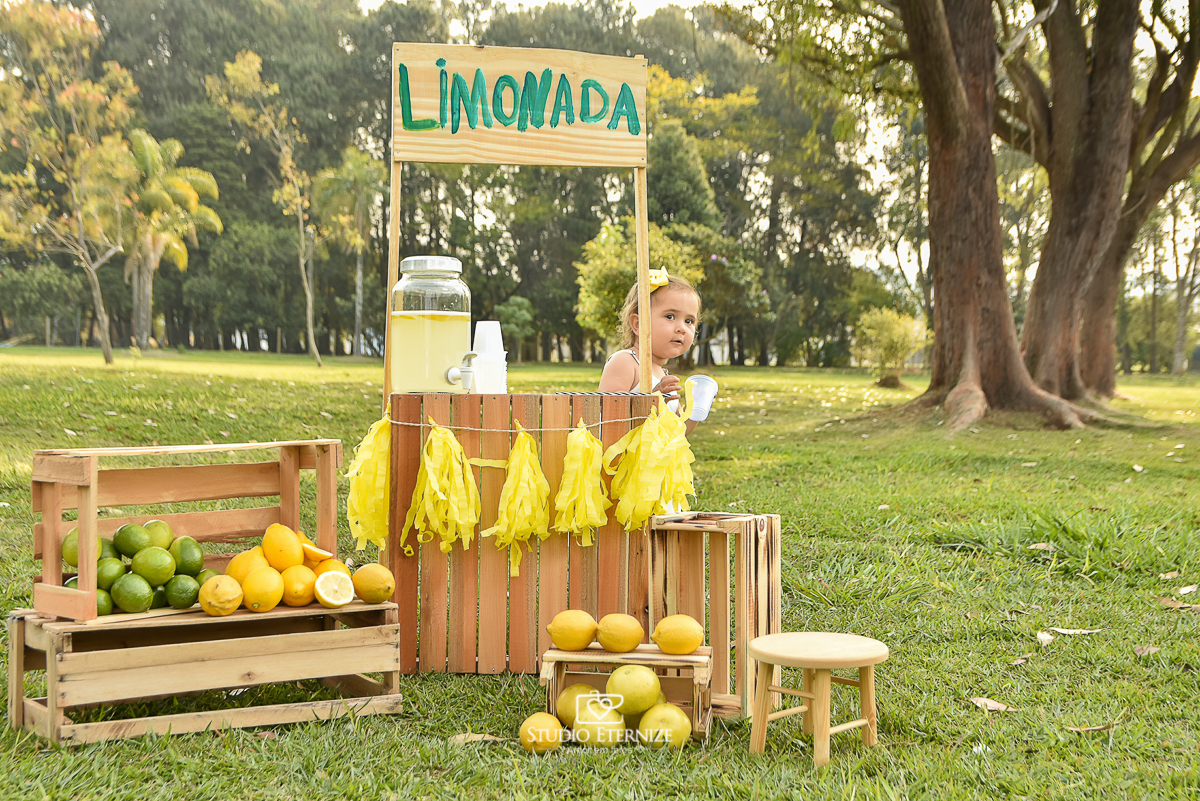 Sessão fotográfica infantil externa com cenário da barraca da limonada. A criança estava atrás da barraca segurando um copo de suco.