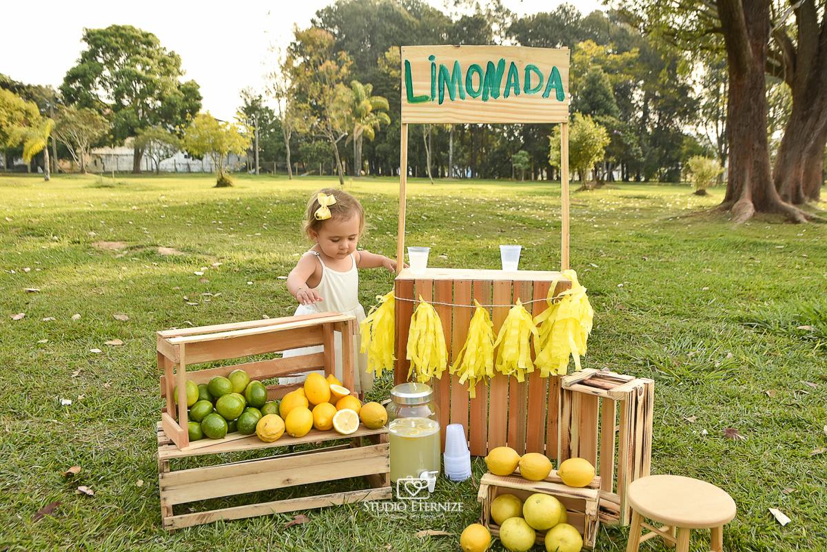 Sessão fotográfica infantil externa com cenário da barraca da limonada. A criança estava atrás da barraca solhando para os limões que estão decorando o chão.