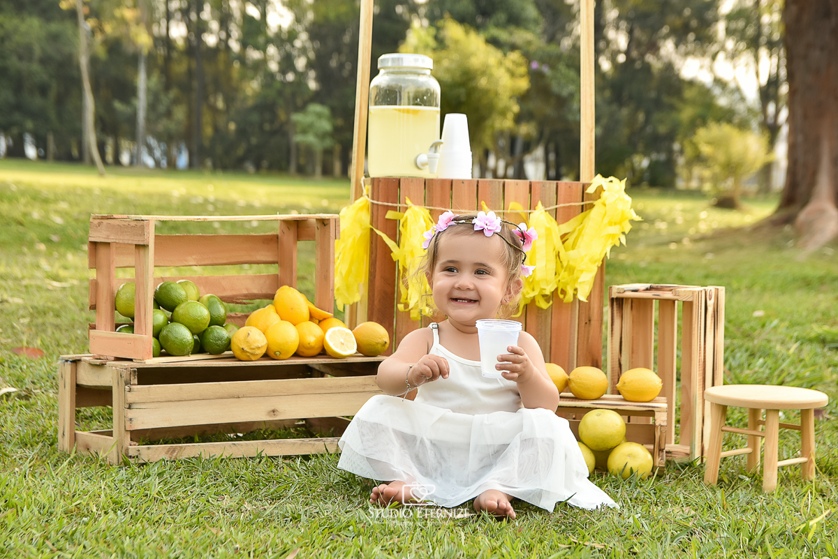 Sessão fotográfica infantil externa com cenário da barraca da limonada. A criança está sentada na frente da 
 barraca da limonada segurando um copo de suco e sorrindo.