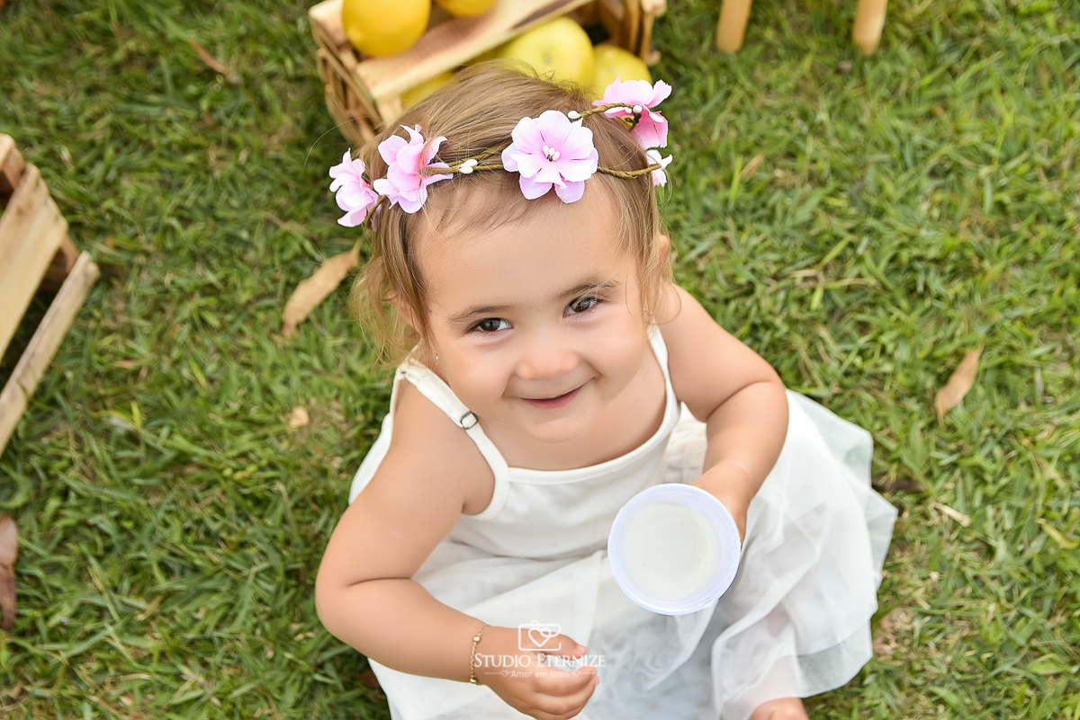 Sessão fotográfica infantil externa com cenário da barraca da limonada. A criança está sentada na frente da 
 barraca da limonada segurando um copo de suco e sorrindo, close de cima.