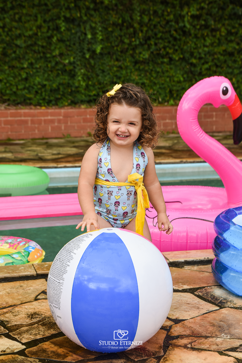 Ensaio fotográfico infantil de verão realizado pelo Studio Eternize em Poços de Caldas - MG. Cenário montado na beira da piscina, com cadeira de praia e boias. A criança está em pé em frente ao cenário segurando uma bola de piscina.