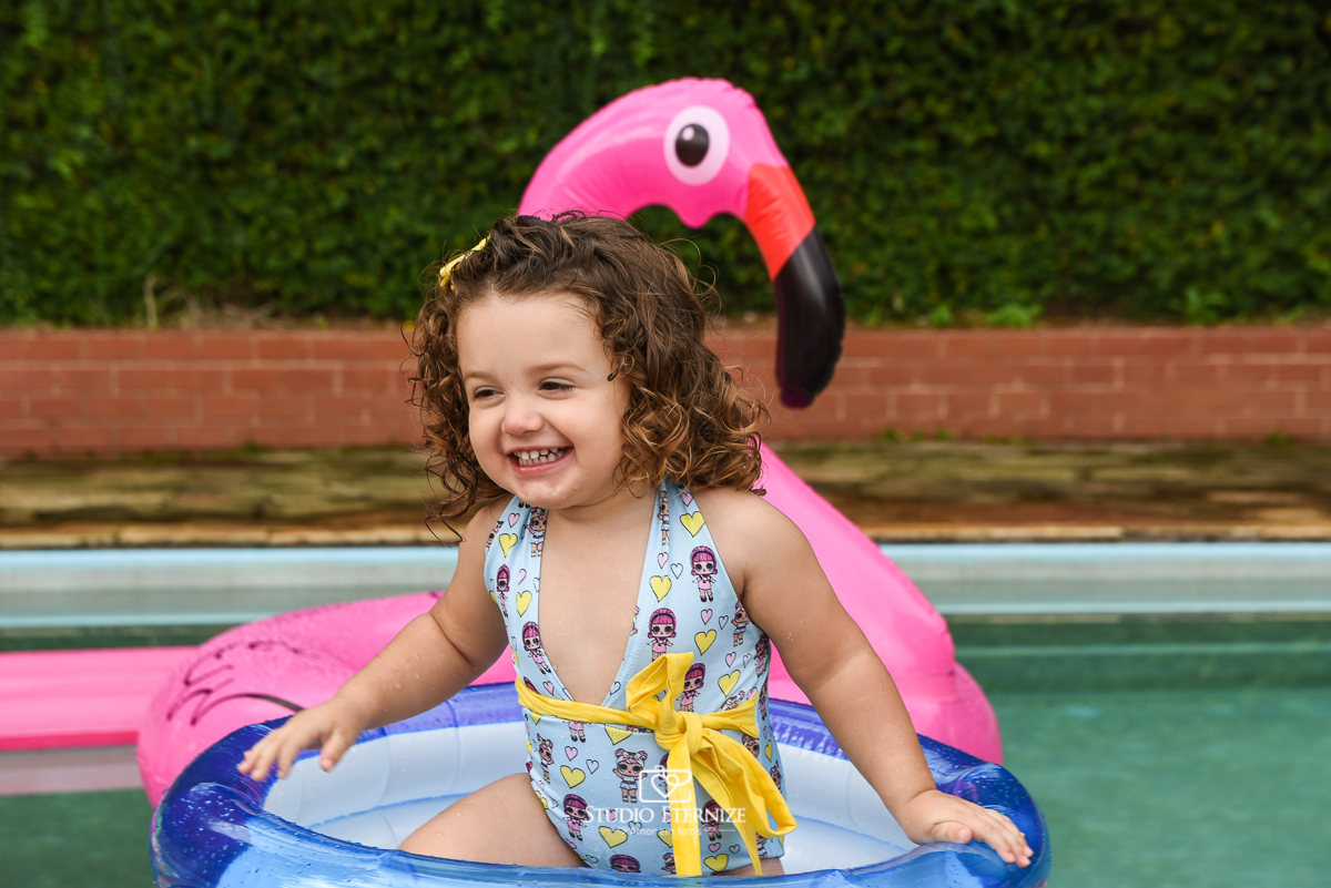 Ensaio fotográfico infantil de verão realizado pelo Studio Eternize em Poços de Caldas - MG. Cenário montado na beira da piscina, com cadeira de praia e boias. A criança está em uma piscina inflável se divertindo.