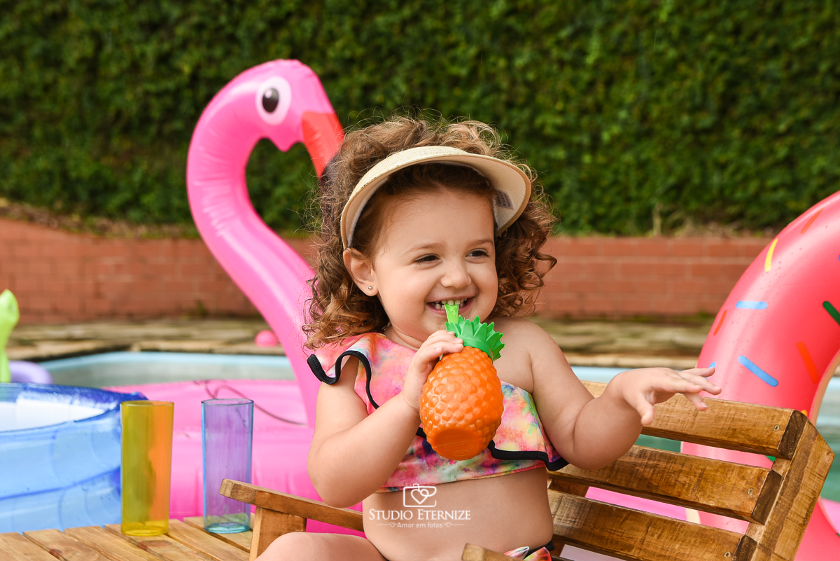 Ensaio fotográfico infantil de verão realizado pelo Studio Eternize em Poços de Caldas - MG. Cenário montado na beira da piscina, com cadeira de praia e boias. Close da criança sentada na cadeira de praia bebendo suco em um copo de abacaxi.