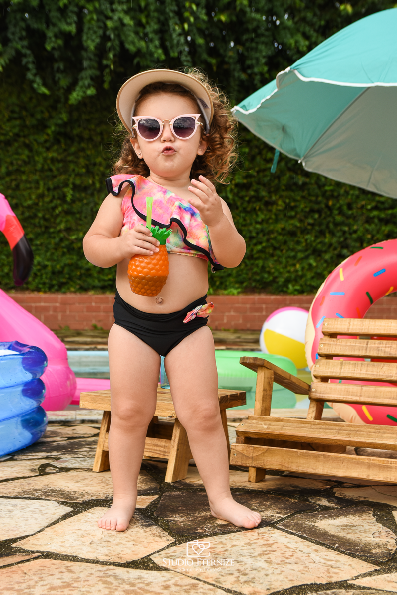 Ensaio fotográfico infantil de verão realizado pelo Studio Eternize em Poços de Caldas - MG. Cenário montado na beira da piscina, com cadeira de praia e boias. A criança está em pé em frente a cadeira de praia segurando um copo de abacaxi e de óculos.