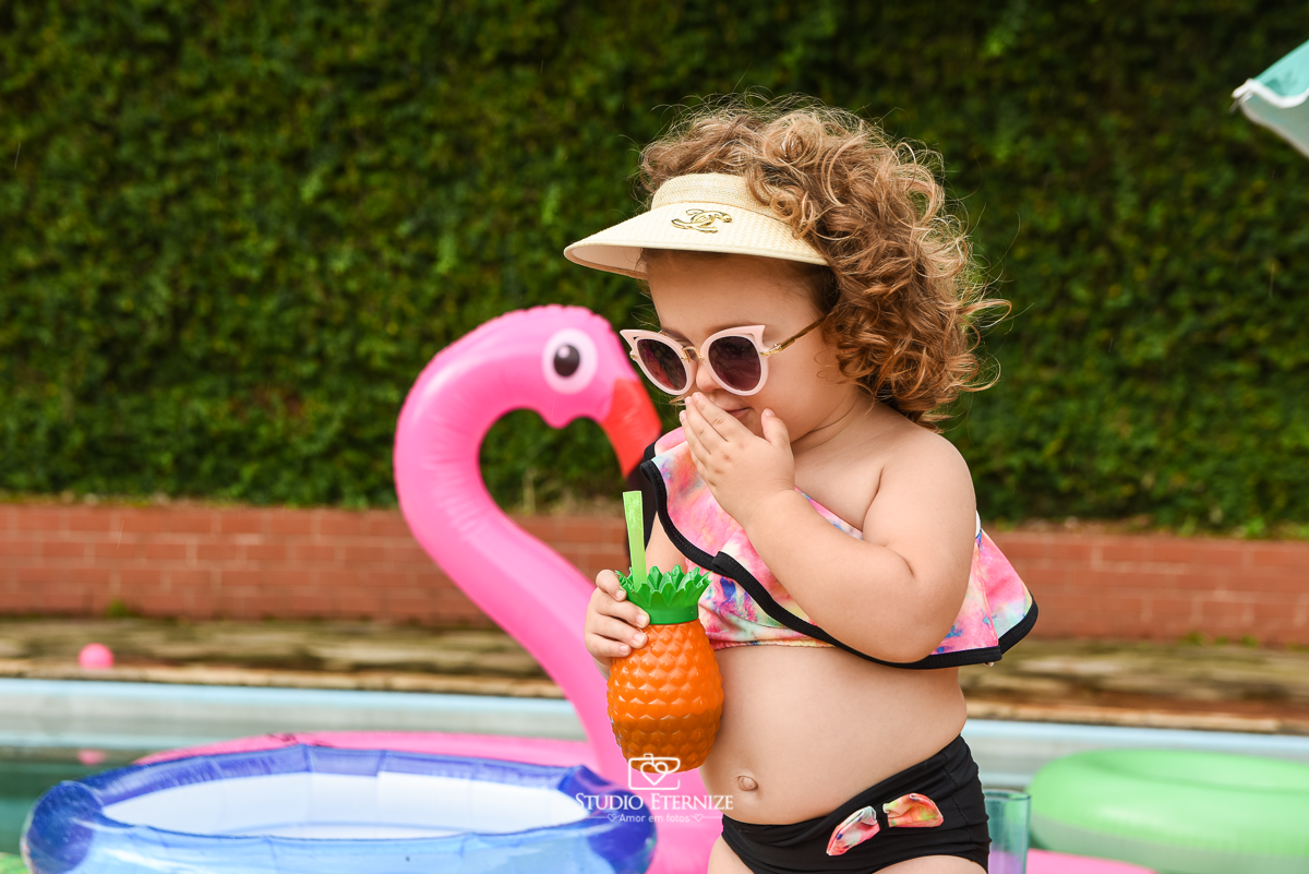 Ensaio fotográfico infantil de verão realizado pelo Studio Eternize em Poços de Caldas - MG. Cenário montado na beira da piscina, com cadeira de praia e boias. Close da criança em frente a cadeira de praia com um copo de abacaxi e arrumando seu óculos.