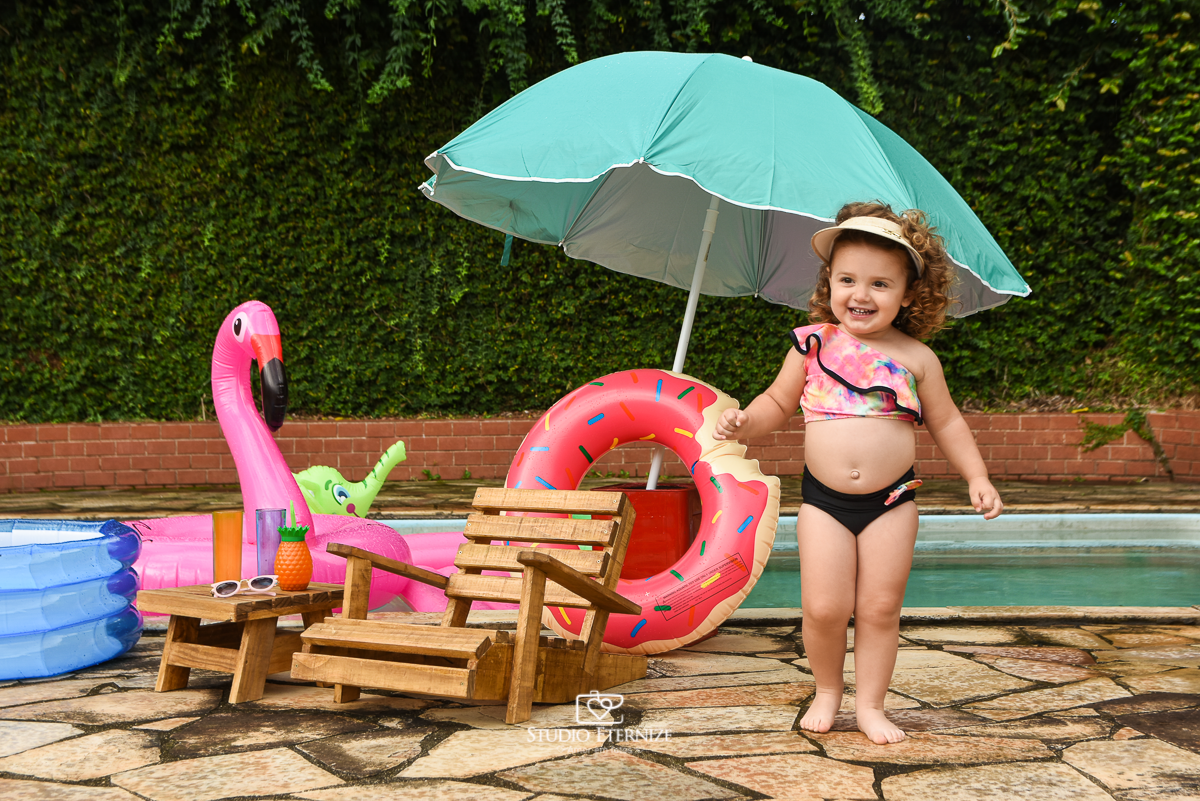 Ensaio fotográfico infantil de verão realizado pelo Studio Eternize em Poços de Caldas - MG. Cenário montado na beira da piscina, com cadeira de praia e boias. A criança está em pé em frente a cadeira de praia.