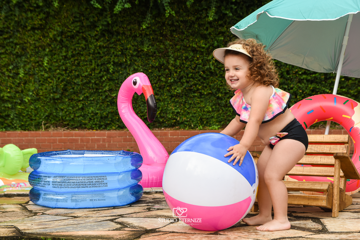 Ensaio fotográfico infantil de verão realizado pelo Studio Eternize em Poços de Caldas - MG. Cenário montado na beira da piscina, com cadeira de praia e boias. A criança está em pé em frente a cadeira de piscina segurando uma bola de piscina.
