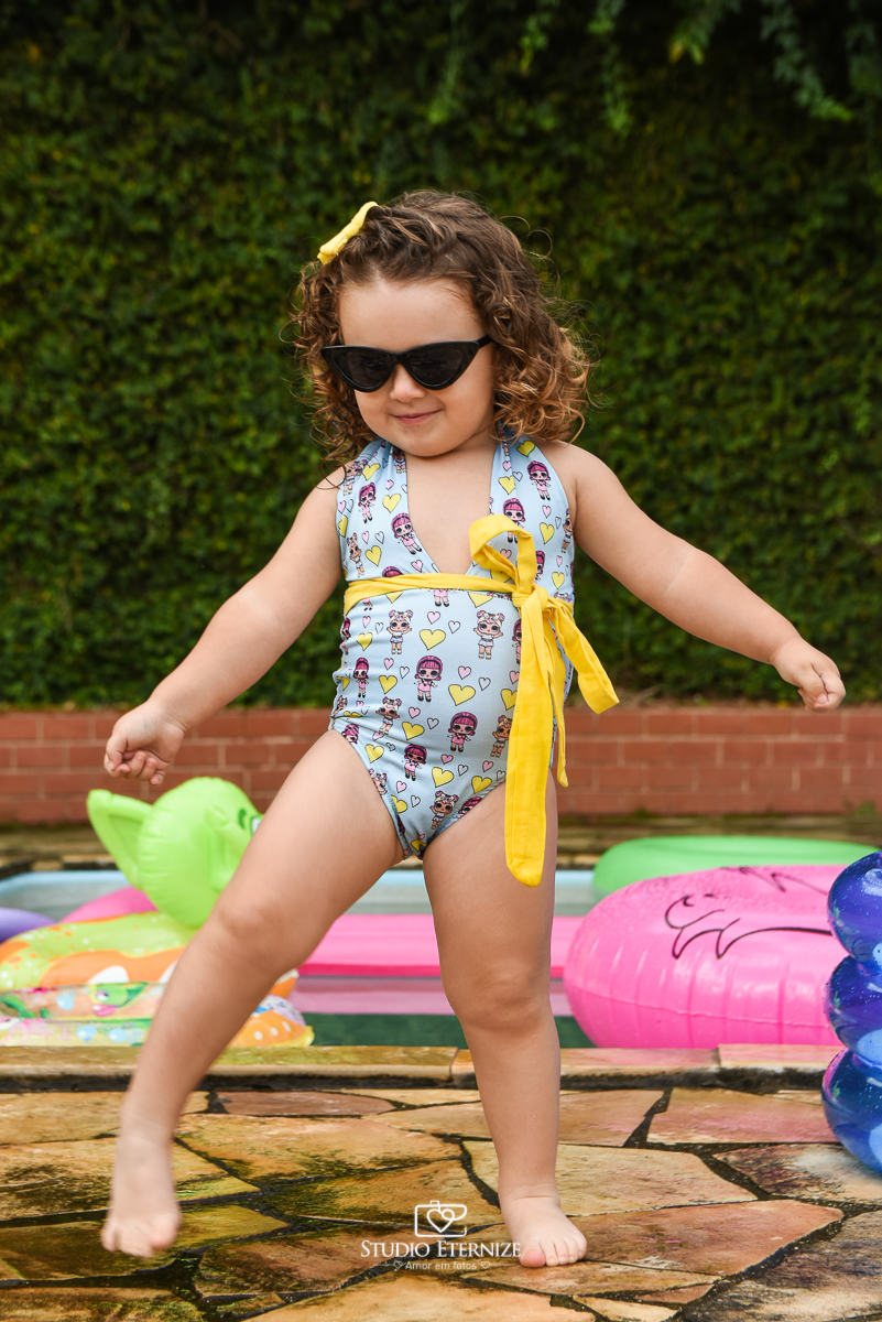 Ensaio fotográfico infantil de verão realizado pelo Studio Eternize em Poços de Caldas - MG. Cenário montado na beira da piscina, com cadeira de piscina e boias. A criança está em pé em frente o cenário de óculos de sol fazendo pose.