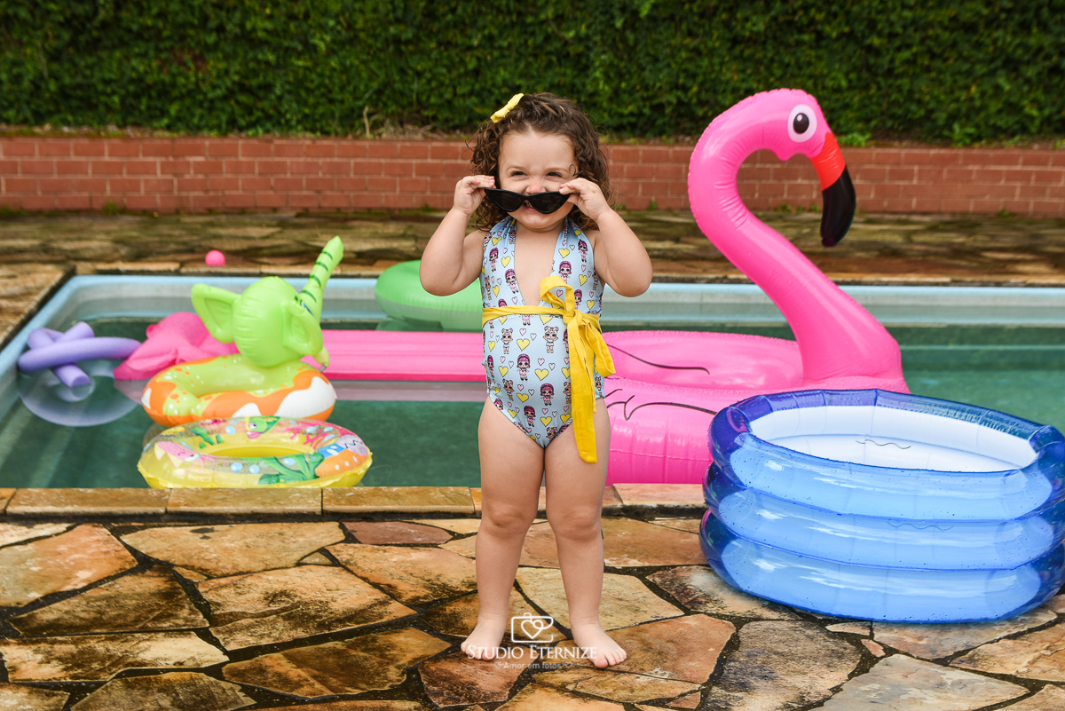Ensaio fotográfico infantil de verão realizado pelo Studio Eternize em Poços de Caldas - MG. Cenário montado na beira da piscina, com cadeira de praia e boias. A criança está em pé em frente ao cenário arrumando seu óculos.
