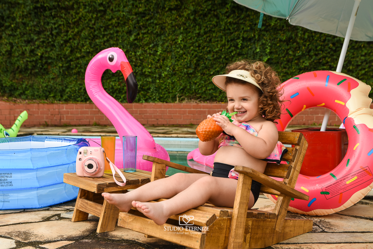 Ensaio fotográfico infantil de verão realizado pelo Studio Eternize em Poços de Caldas - MG. Cenário montado na beira da piscina, com cadeira de praia e boias. A criança está sentada na cadeira de praia bebendo suco em um copo de abacaxi.