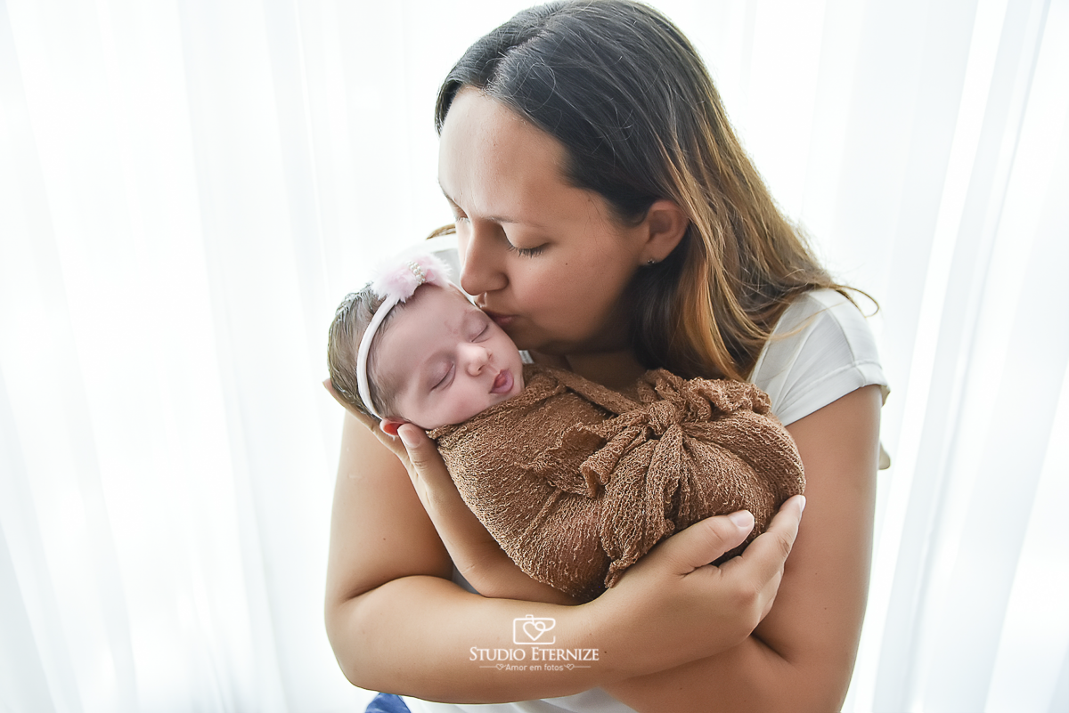 Fotografia newborn menina realizada pelo Studio Eternize em Poços de Caldas - MG. Bebê no colo da mãe enroladinha no wrap. Mãe dando um beijo em seu rosto.