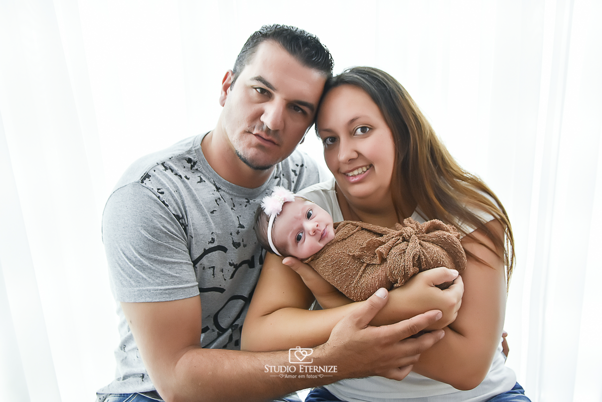 Fotografia newborn menina realizada pelo Studio Eternize em Poços de Caldas - MG. Bebê no colo da mãe enroladinha no wrap e de olhos abertos. Pai está ao lado da mãe abraçando-a.
