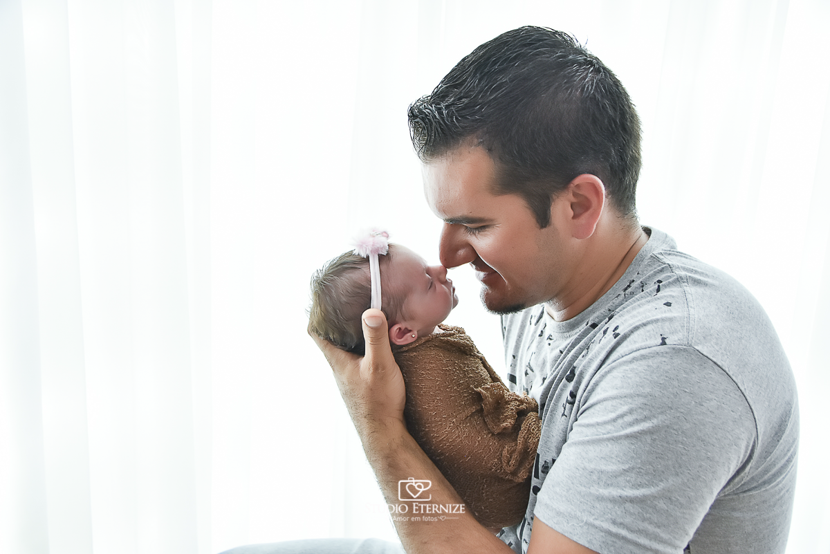 Fotografia newborn menina realizada pelo Studio Eternize em Poços de Caldas - MG. Bebê no colo do pai, de frente para ele, enroladinha no wrap. Pai encosta seu nariz no da filha. Beijinho de esquimó.