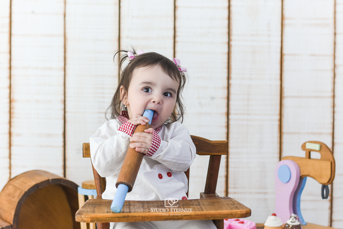Fotografia de acompanhamento mensal do bebê realizada no Studio Eternize em Poços de Caldas - MG. Bebê menina com 9 meses de idade, tema da sessão de cozinheiro. Ela está vestida de cozinheira, sentada no cadeirão. mordendo o rolo de macarrão.