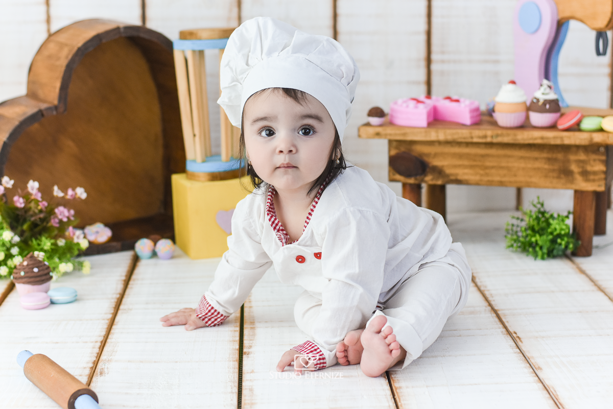 Fotografia de acompanhamento mensal do bebê realizada no Studio Eternize em Poços de Caldas - MG. Bebê menina com 9 meses de idade, tema da sessão de cozinheiro. Ela está vestida de cozinheira, em frente ao cenário.