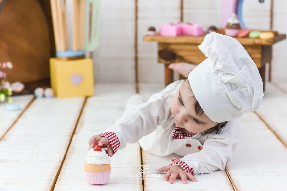 Fotografia de acompanhamento mensal do bebê realizada no Studio Eternize em Poços de Caldas - MG. Bebê menina com 9 meses de idade, tema da sessão de cozinheiro. Ela está vestida de cozinheira, em frente ao cenário, de bruços, tentando pegar um cupcake.