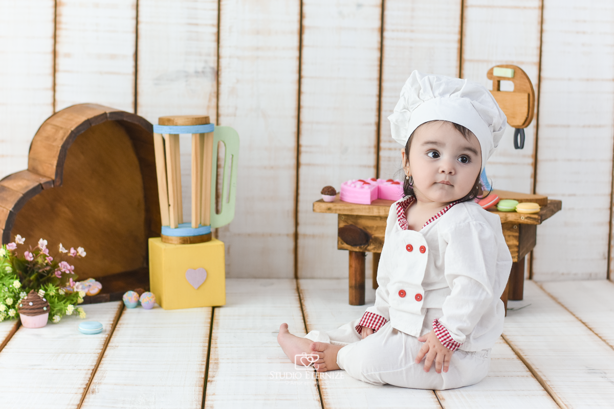 Fotografia de acompanhamento mensal do bebê realizada no Studio Eternize em Poços de Caldas - MG. Bebê menina com 9 meses de idade, tema da sessão de cozinheiro. Ela está vestida de cozinheira, sentada de lado no cenário.