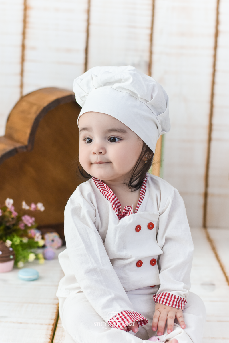 Fotografia de acompanhamento mensal do bebê realizada no Studio Eternize em Poços de Caldas - MG. Bebê menina com 9 meses de idade, tema da sessão de cozinheiro. Ela está vestida de cozinheira, sentada no cenário olhando para o lado.
