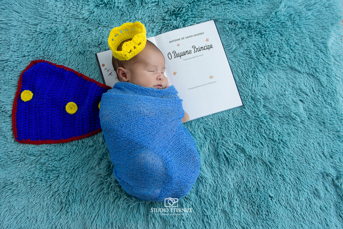 Fotografia newborn menino realizada pelo Studio Eternize em Poços de Caldas - MG. Bebê enrolado em um wrap azul, com coroa e capa de príncipe, deitado de lado no livro do Pequeno Príncipe.