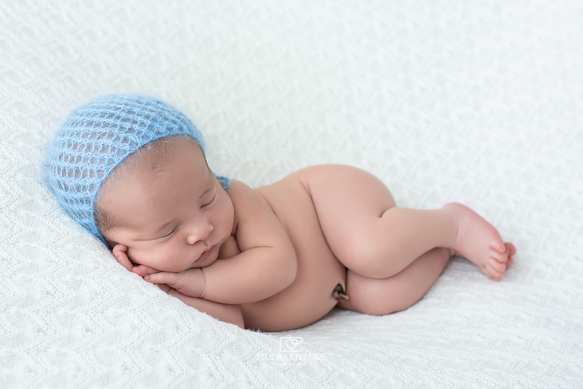 Fotografia newborn de menino realizada em Poços de Caldas - MG, pelo Studio Eternize Amor em Fotos. Bebê peladinho de touquinha azul deitado de ladinho.