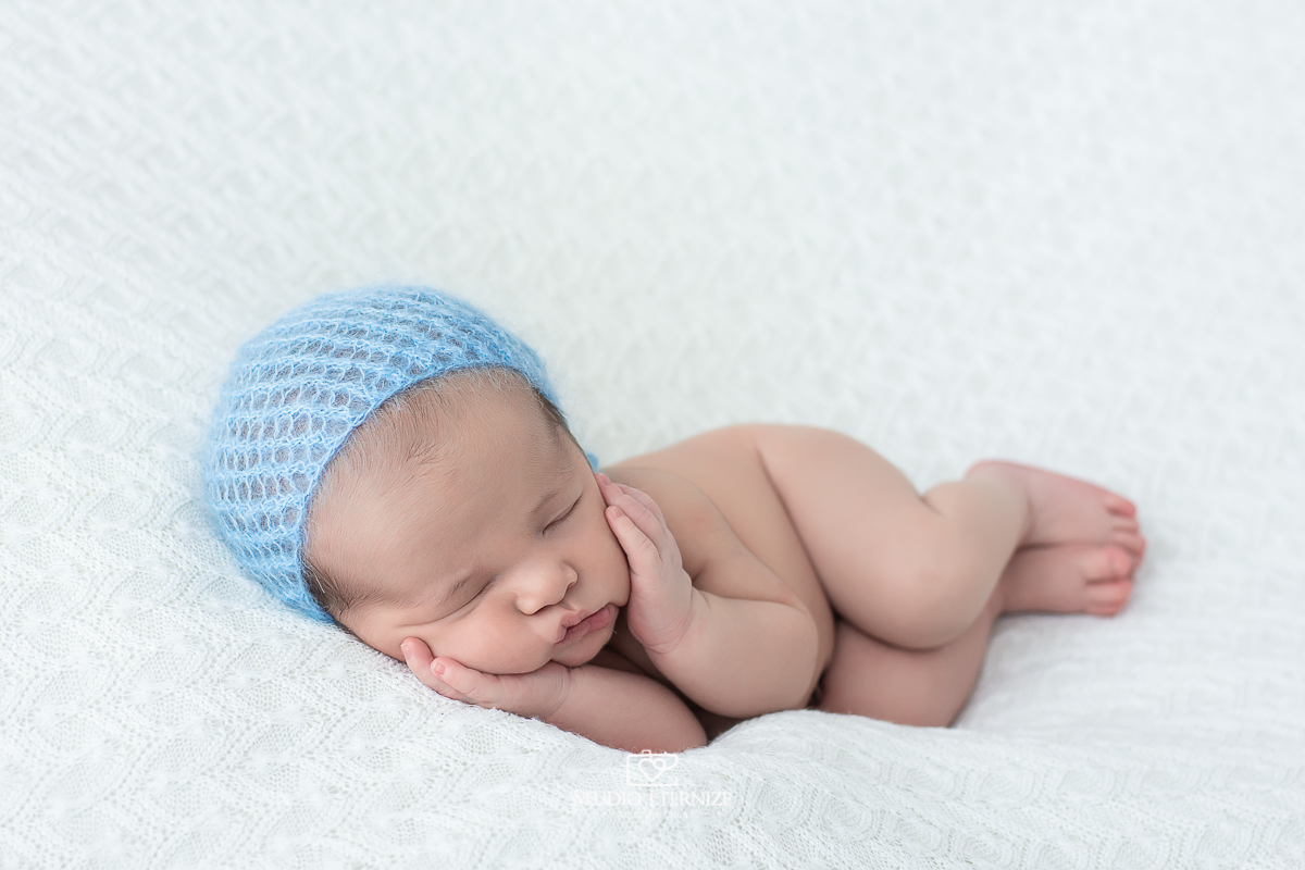 Fotografia newborn menino realizada pelo Studio Eternize Amor em fotos, em Poços de Caldas - MG. Bebê peladinho de touquinha azul deitado de ladinho com as duas mãos na bochecha.