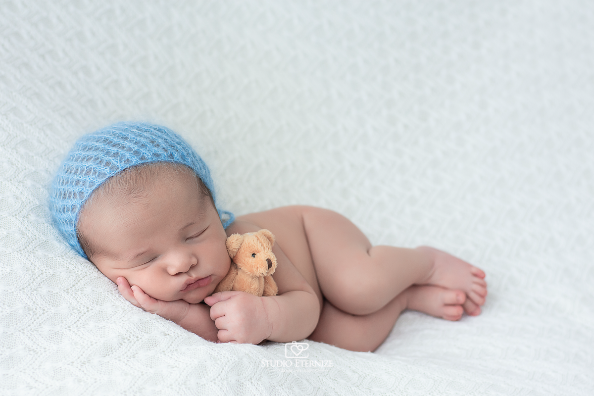 Fotografia newborn menino realizada pelo Studio Eternize Amor em fotos, na cidade de Poços de Caldas - MG. Bebê peladinho de touquinha azul deitado de ladinho com ursinho.